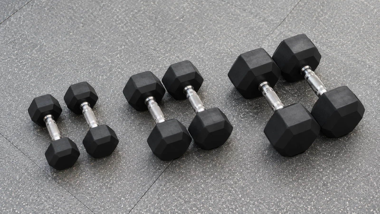 Three pairs of black hex dumbbells in increasing sizes arranged on a gym floor.