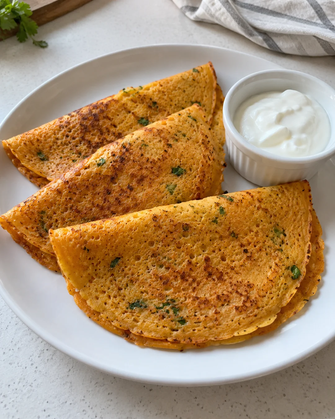Thin savory red lentil pancakes folded on a white plate with yogurt on the side.